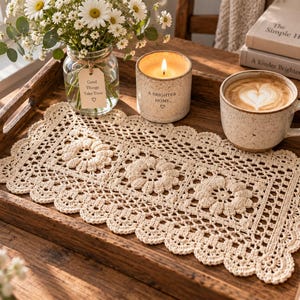 Puede incluir: Un tapete de ganchillo rectangular de color crema con tres motivos florales centrales y un borde festoneado. El tapete está colocado sobre una bandeja de madera marrón oscuro junto a un jarrón de cristal con flores blancas, una vela y una taza.