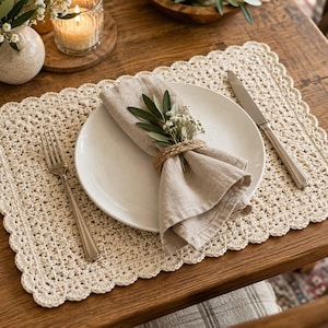 May include: A rectangular cream-colored crocheted placemat with a scalloped edge, featuring a white ceramic plate, a beige linen napkin held by a braided rope ring, a silver fork, and a silver knife arranged on a wooden surface.