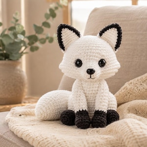 May include: A handcrafted crochet fox toy featuring a white body, a large bushy tail, and dark brown accents on the tips of the ears and paws. The fox has large, round, glossy black eyes and a small black nose, sitting in an upright position.