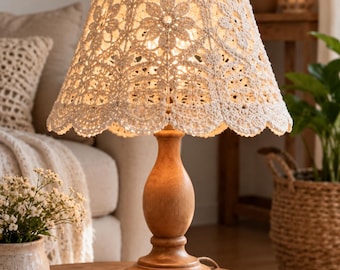 May include: A table lamp with a light wood base and a cream-colored crocheted lampshade featuring a floral pattern. The shade has a scalloped edge and emits a warm glow from the light source inside, illuminating the intricate yarn work.