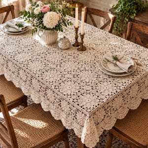May include: A rectangular beige crochet tablecloth featuring a repeating square floral motif pattern with a scalloped edge. The tablecloth is draped over a wooden dining table, providing a textured surface for place settings and a central floral arrangement.