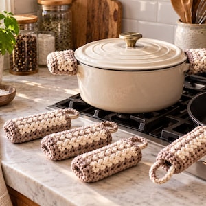 May include: A set of crochet handle covers featuring a two-tone pattern of cream and light brown yarn. The cylindrical covers have a textured, braided stitch design and include a small loop at one end for hanging, designed to fit cookware handles.