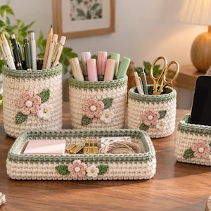 May include: A set of five cream-colored crochet desk organizers with sage green trim and floral appliques. The set includes three cylindrical pen holders, one rectangular tray, and one square holder containing a black smartphone.