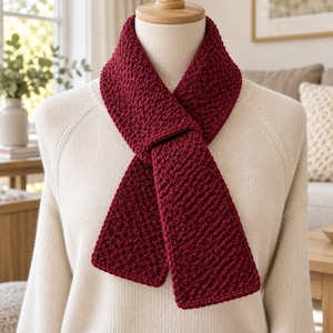 May include: A deep red knitted scarf with a textured seed stitch pattern, draped around the neck of a mannequin wearing a cream-colored long-sleeve sweater. The scarf features a consistent, dense knit construction with a rectangular shape.