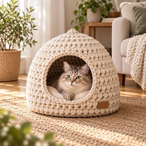 May include: A cream-colored, chunky knit cat bed with a dome shape and a wide circular opening. A long-haired tabby cat with green eyes is resting inside the soft, textured structure. A small, rectangular brown leather-like tag is attached to the front.