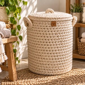 May include: A cylindrical, cream-colored crochet laundry basket with a matching lid and two side handles. The basket features a small, rectangular brown leather-like tag on the front with the word "Home" and a small heart symbol printed on it.