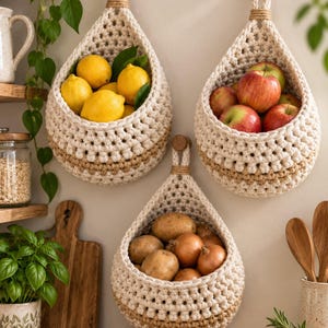 May include: Three teardrop-shaped hanging baskets made of off-white and tan woven cord. Each basket is suspended from a wooden wall hook and contains fresh produce: one holds yellow lemons, one holds red apples, and one holds potatoes and onions.