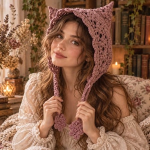 May include: A dusty rose crochet bonnet featuring a pointed cat-ear silhouette and an openwork lace pattern. The headpiece includes two long braided ties that hang down, each finished with a decorative crocheted flower at the bottom end.