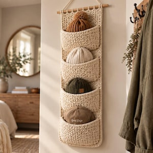 Peut inclure: Un organisateur mural vertical en crochet de couleur crème doté de quatre poches profondes. Chaque poche contient un bonnet différent : un bonnet à pompon marron, un bonnet côtelé crème, un bonnet vert foncé et une casquette beige Good Days.