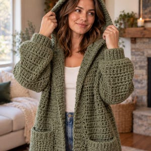 Può includere: Un cardigan con cappuccio color verde salvia realizzato in filato di uncinetto spesso e voluminoso. Il capo presenta maniche lunghe con polsini a coste, due ampie tasche frontali applicate e un design aperto sul davanti senza chiusure.