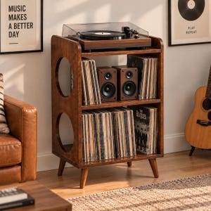 May include: A brown wooden record player stand with two circular side cutouts, four tapered legs, and two shelves. The top shelf holds a black turntable, while the shelves are filled with vinyl records and two black speakers placed in the center.