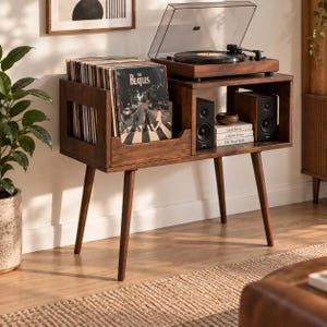 May include: A dark wood record player console with four tapered legs, featuring a dedicated storage section for vinyl records, a top surface for a turntable, and an open shelf housing two black speakers and a stack of three books.