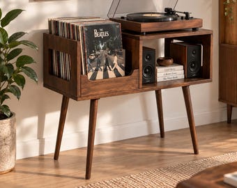 May include: A dark wood record player console with four tapered legs, featuring a dedicated storage section for vinyl records, a top surface for a turntable, and an open shelf housing two black speakers and a stack of three books.