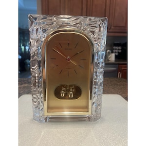 May include: A rectangular Seiko quartz mantel clock featuring a clear, textured glass frame with an arched top. The clock face is gold-toned with thin black hour markers and hands. A gold pendulum with two small weights hangs in the lower section.