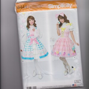 May include: A Simplicity 8444 sewing pattern envelope featuring two dress designs. One dress is blue and white checkered with a pink bow, and the other is pink with a yellow bow and macaron print. Both include white blouses and white shoes.