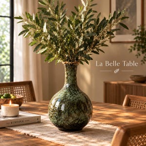 Può includere: Un vaso in ceramica con base bulbosa e collo stretto, caratterizzato da una smaltatura maculata verde e marrone scuro. Il vaso è riempito con rami verdi frondosi. Il testo "La Belle Table" è visibile nell'angolo in alto a destra dell'immagine.