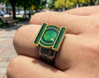 May include: A ring featuring a gold-toned rectangular frame with a central circular emblem, both filled with a textured, glittery green inlay. The band of the ring is a smooth, dark metallic gray, contrasting with the bright green and gold top.