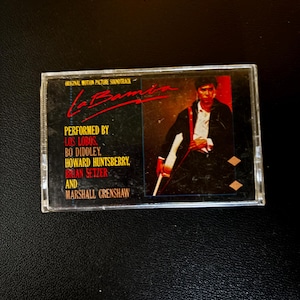 May include: A La Bamba original motion picture soundtrack cassette tape. The black cover features red text, a photograph of a musician holding a guitar, and a list of performers including Los Lobos, Bo Diddley, Howard Huntsberry, Brian Setzer, and Marshall Crenshaw.