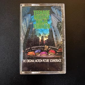 May include: A clear plastic cassette tape case featuring the Teenage Mutant Ninja Turtles The Original Motion Picture Soundtrack cover art. The image displays four turtles with colored masks emerging from a manhole cover in a city.