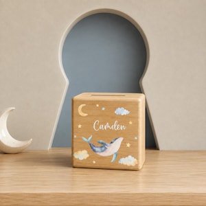 Può includere: Un salvadanaio quadrato in legno con un'illustrazione stampata di una balena blu, una luna crescente gialla e nuvole. Il nome Camden è stampato in corsivo bianco al centro. La scatola presenta una fessura sulla parte superiore per le monete.