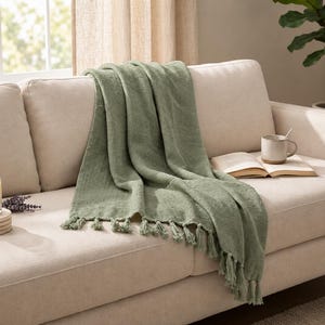 May include: A sage green throw blanket with a textured weave and knotted fringe trim draped over a beige sofa. The blanket features a soft, matte finish and hangs in loose folds, showcasing the detailed fringe along the bottom edge.