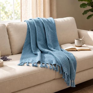 May include: A textured blue throw blanket with knotted fringe tassels draped over a beige sofa. The blanket features a woven fabric construction with a consistent solid color throughout, showcasing a soft appearance and decorative edge detailing.