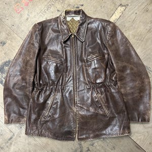 May include: A dark brown leather jacket featuring a front zip fastening, a pointed collar, and two zipped chest pockets. The waist is cinched with an elasticated band, and the interior lining displays a tan and brown plaid pattern.