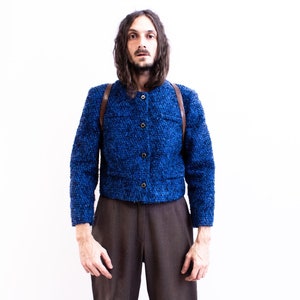 May include: A blue tweed jacket with a brown leather strap. The jacket has a button closure and is worn over a brown pair of trousers.