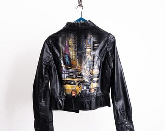 Könnte beinhalten: Schwarze Lederjacke mit einem farbenfrohen Stadtlandschaftsdesign auf dem Rücken. Das Kunstwerk zeigt eine Nachtszene mit Gebäuden, Lichtern und gelben Taxis. Die Jacke hat lange Ärmel und geknöpfte Manschetten.