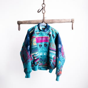 Könnte beinhalten: Ein türkisfarbener Vintage-Pullover mit einem bunten geometrischen Muster und dem Wort "TAURUS" auf der Brust. Der Pullover hat einen blauen Kragen und Bündchen und hängt an einem Holzgestell.