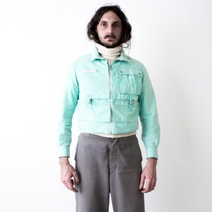 May include: A mint green cropped jacket featuring a full-length silver zipper, a high collar, and multiple utility pockets on the front. The jacket is worn over a cream-colored turtleneck sweater and paired with plain gray trousers.