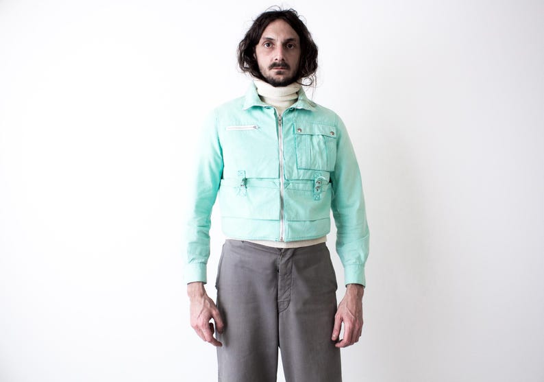 May include: A mint green cropped jacket featuring a full-length silver zipper, a high collar, and multiple utility pockets on the front. The jacket is worn over a cream-colored turtleneck sweater and paired with plain gray trousers.