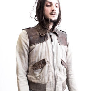 May include: A man wearing a light brown leather and beige jacket with a zipper closure. The jacket has a brown leather panel on the shoulders and a brown leather pocket on the left side.