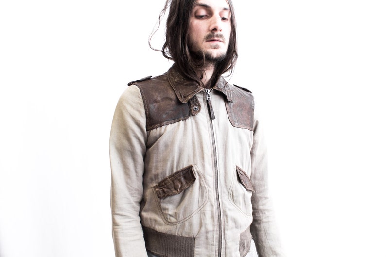 May include: A man wearing a light brown leather and beige jacket with a zipper closure. The jacket has a brown leather panel on the shoulders and a brown leather pocket on the left side.