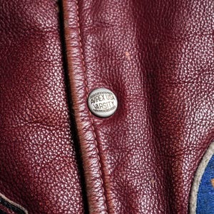 May include: A deep burgundy leather jacket featuring a silver-toned metal snap button embossed with the text AVIREX USA VARSITY. The leather has a visible pebbled texture, and a portion of a blue and gold embroidered patch is visible on the right.
