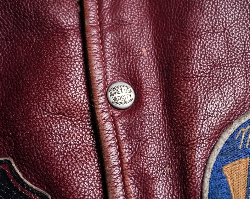 May include: A deep burgundy leather jacket featuring a silver-toned metal snap button embossed with the text AVIREX USA VARSITY. The leather has a visible pebbled texture, and a portion of a blue and gold embroidered patch is visible on the right.