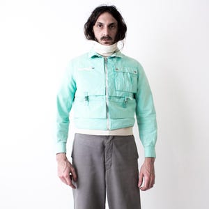 May include: A light turquoise jacket with multiple front pockets and a white zipper. The jacket is paired with gray pants and a white turtleneck. The outfit is a vintage style.
