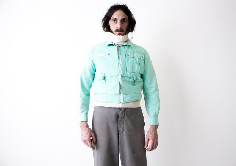 May include: A light turquoise jacket with multiple front pockets and a white zipper. The jacket is paired with gray pants and a white turtleneck. The outfit is a vintage style.