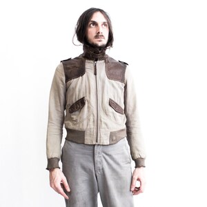 May include: A man wearing a light brown bomber jacket with brown leather accents and gray pants. The jacket has a zipper closure and two pockets on the front.