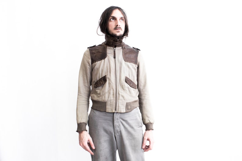 May include: A man wearing a light brown bomber jacket with brown leather accents and gray pants. The jacket has a zipper closure and two pockets on the front.
