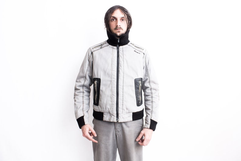 K&ouml;nnte beinhalten: Eine graue Bomberjacke mit schwarzem Besatz und Rei&szlig;verschlusstaschen. Die Jacke hat einen hohen Kragen und einen Rei&szlig;verschluss.