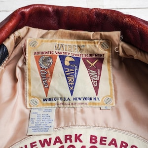 May include: A red and black leather jacket interior featuring a beige lining and a large rectangular label. The label displays the text AVIREX AUTHENTIC VARSITY SPORTS EQUIPMENT with three pennants and a smaller tag detailing material composition.