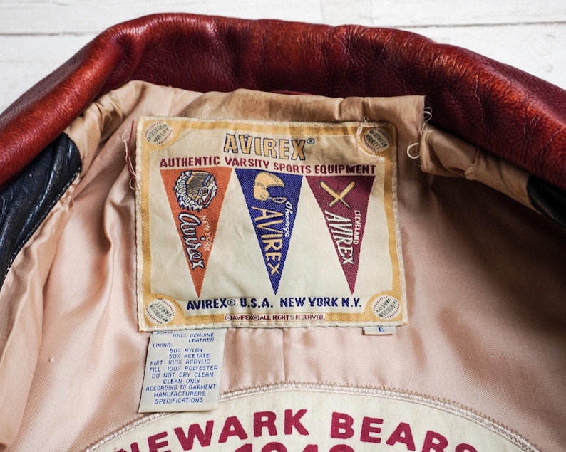 May include: A red and black leather jacket interior featuring a beige lining and a large rectangular label. The label displays the text AVIREX AUTHENTIC VARSITY SPORTS EQUIPMENT with three pennants and a smaller tag detailing material composition.