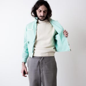 May include: A mint green zip-up jacket with a high collar and a horizontal zippered chest pocket, worn over a cream-colored long-sleeve sweater and paired with grey trousers. The jacket features a front zipper closure and long sleeves.