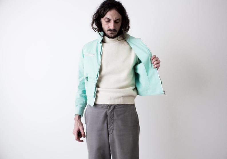 May include: A mint green zip-up jacket with a high collar and a horizontal zippered chest pocket, worn over a cream-colored long-sleeve sweater and paired with grey trousers. The jacket features a front zipper closure and long sleeves.