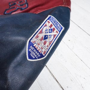 May include: A dark blue and red leather varsity jacket featuring a white embroidered patch on the sleeve. The patch displays the text STATE FINAL CHAMPION, Newark Baseball Team, 1959, and an illustration of crossed baseball bats.