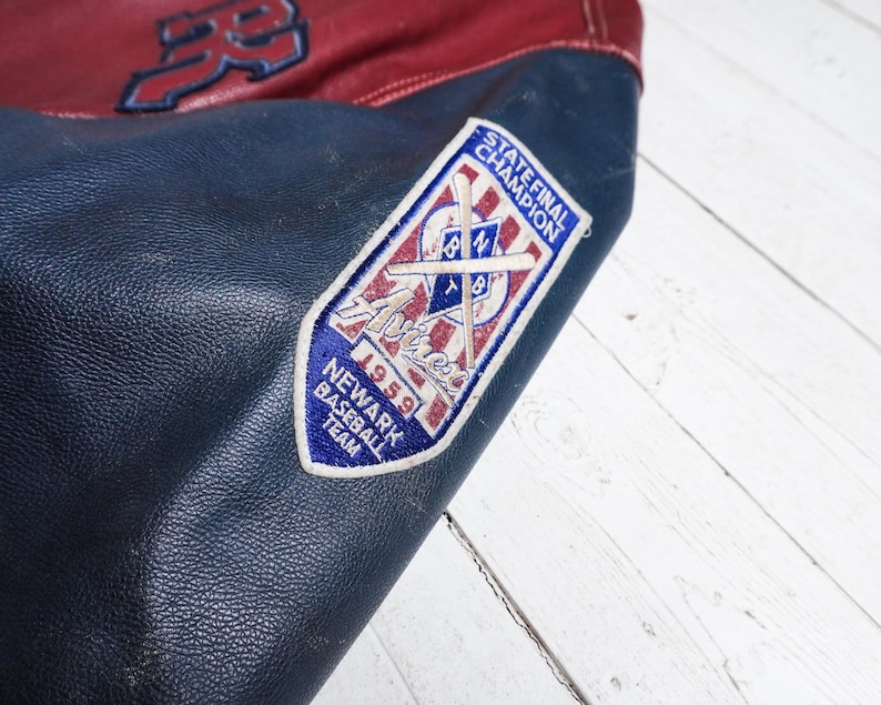 May include: A dark blue and red leather varsity jacket featuring a white embroidered patch on the sleeve. The patch displays the text STATE FINAL CHAMPION, Newark Baseball Team, 1959, and an illustration of crossed baseball bats.