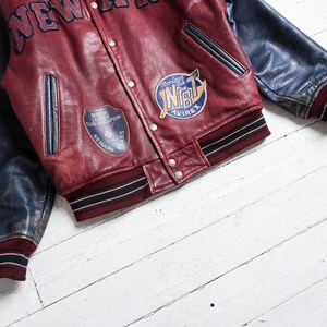 May include: A varsity jacket featuring a deep red leather body and dark blue leather sleeves. The front displays the text NEW YORK, a Newark Bears patch, and an NBT Avirex patch. It includes a snap-button closure, two side pockets, and striped ribbed cuffs and hem.