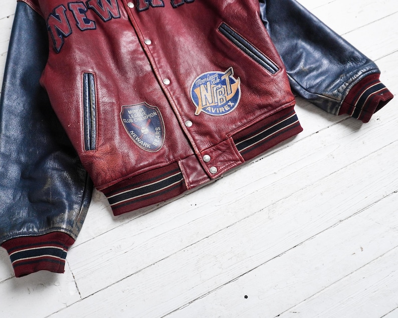 May include: A varsity jacket featuring a deep red leather body and dark blue leather sleeves. The front displays the text NEW YORK, a Newark Bears patch, and an NBT Avirex patch. It includes a snap-button closure, two side pockets, and striped ribbed cuffs and hem.
