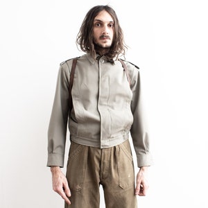 Pu&ograve; includere: Giacca beige chiaro con maniche lunghe, colletto button-down e imbracatura in pelle marrone. La giacca ha un orlo a coste ed &egrave; abbinata a pantaloni marroni. L'outfit &egrave; ambientato su uno sfondo bianco.
